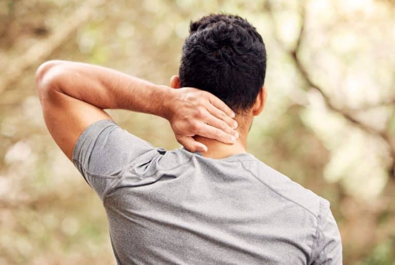 Man in an outdoor setting holding the back of his neck with one hand. The image is from behind and shows the mans back and back of head.