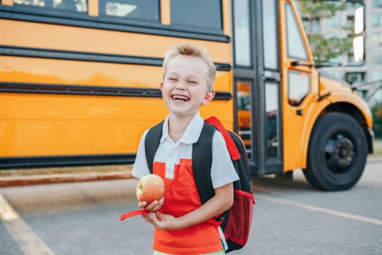 Child with a visible hearing aid standing in front of a school bus. He is holding an apple and has his backpack on. He smiling and mid-laugh.