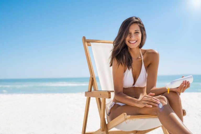 Woman in swimsuit on beach chair at the beach smiling and applying lotion